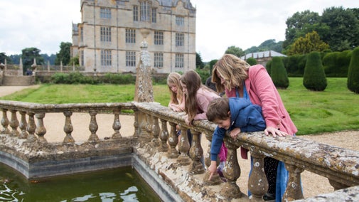 A parent and three children lean over a stone balustrade, pointing at fish in a pond in the formal garden, with Montacute House behind them